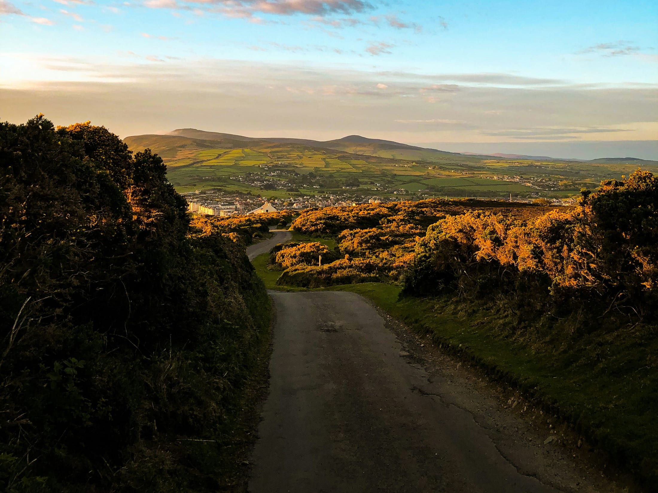 A lane dropping towards a town and the hills of the Isle of Man under a morning sky