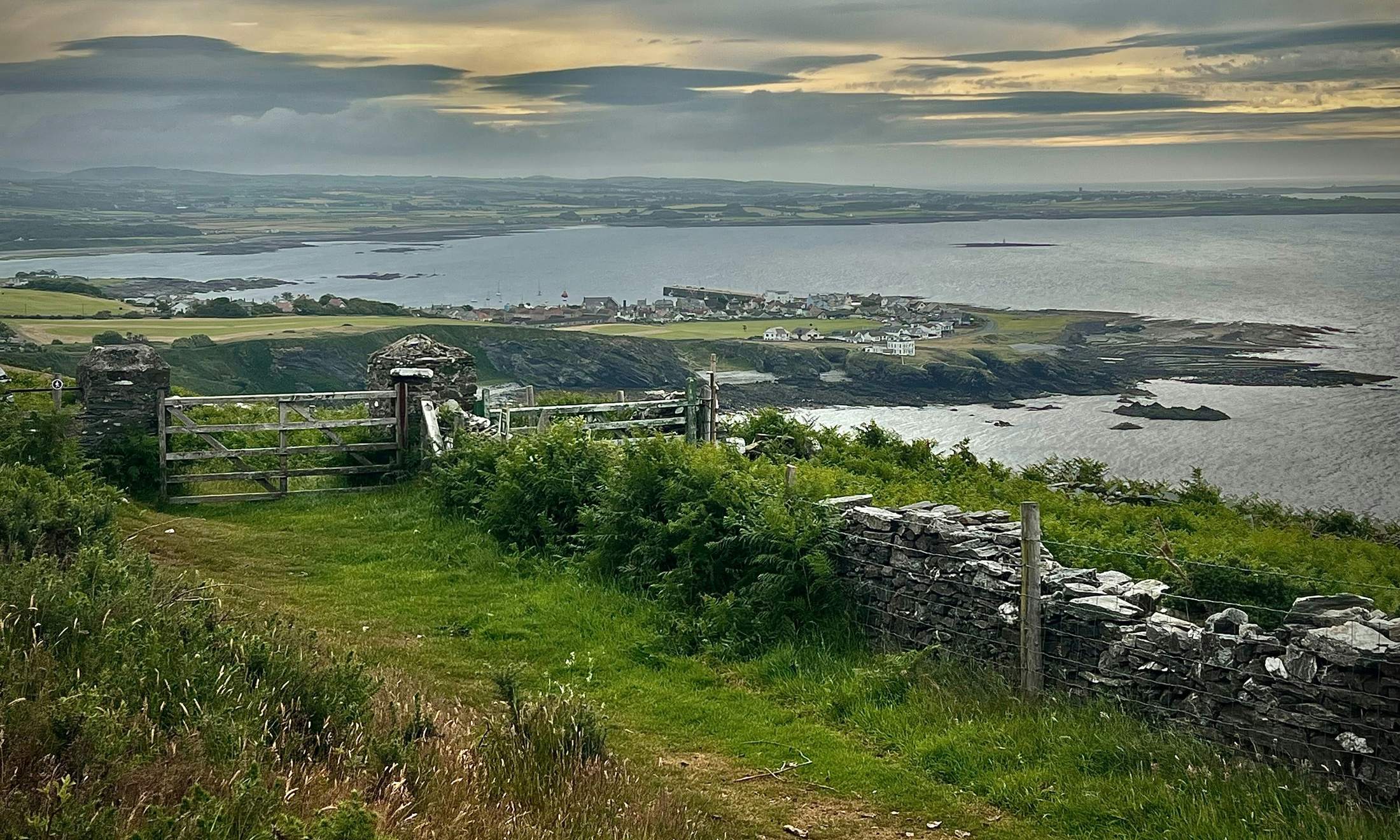 A field gate and grassy track above the coast of the Isle of Man, with a village and the sea beyond under a stormy sky